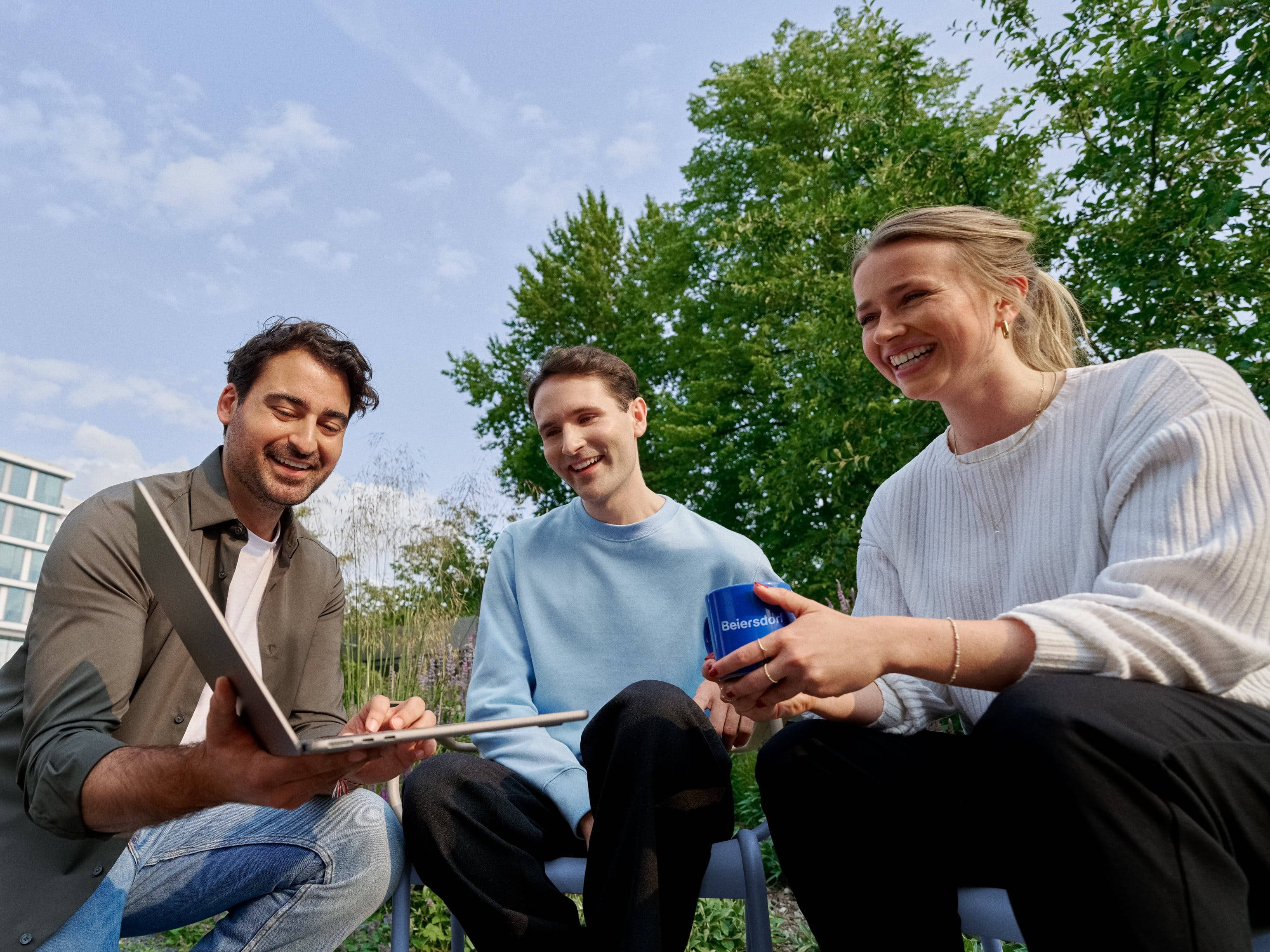 Three smiling coworkers sitting in the campus garden, collaborating on a laptop and enjoying teamwork.