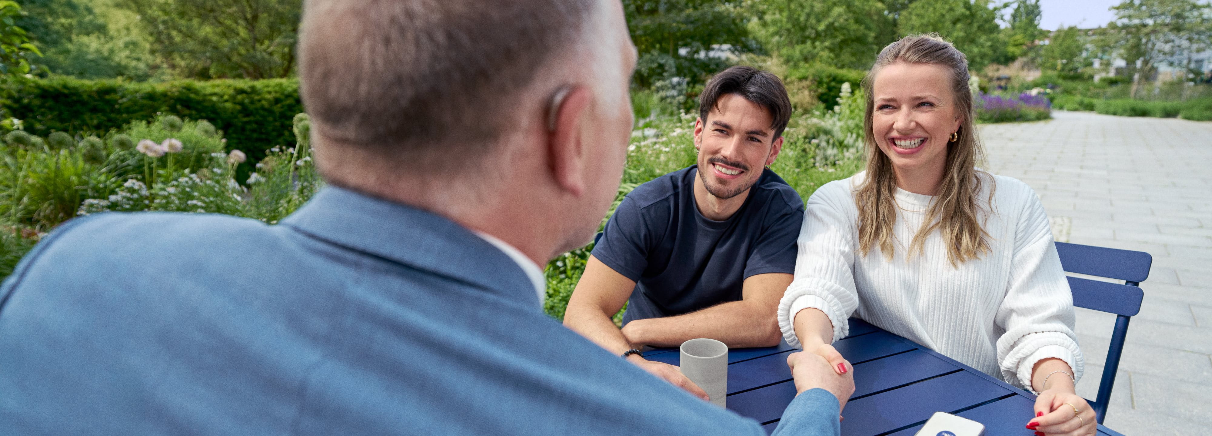 Two colleagues sitting at an outdoor table smiling and talking with a third person during an informal meeting.