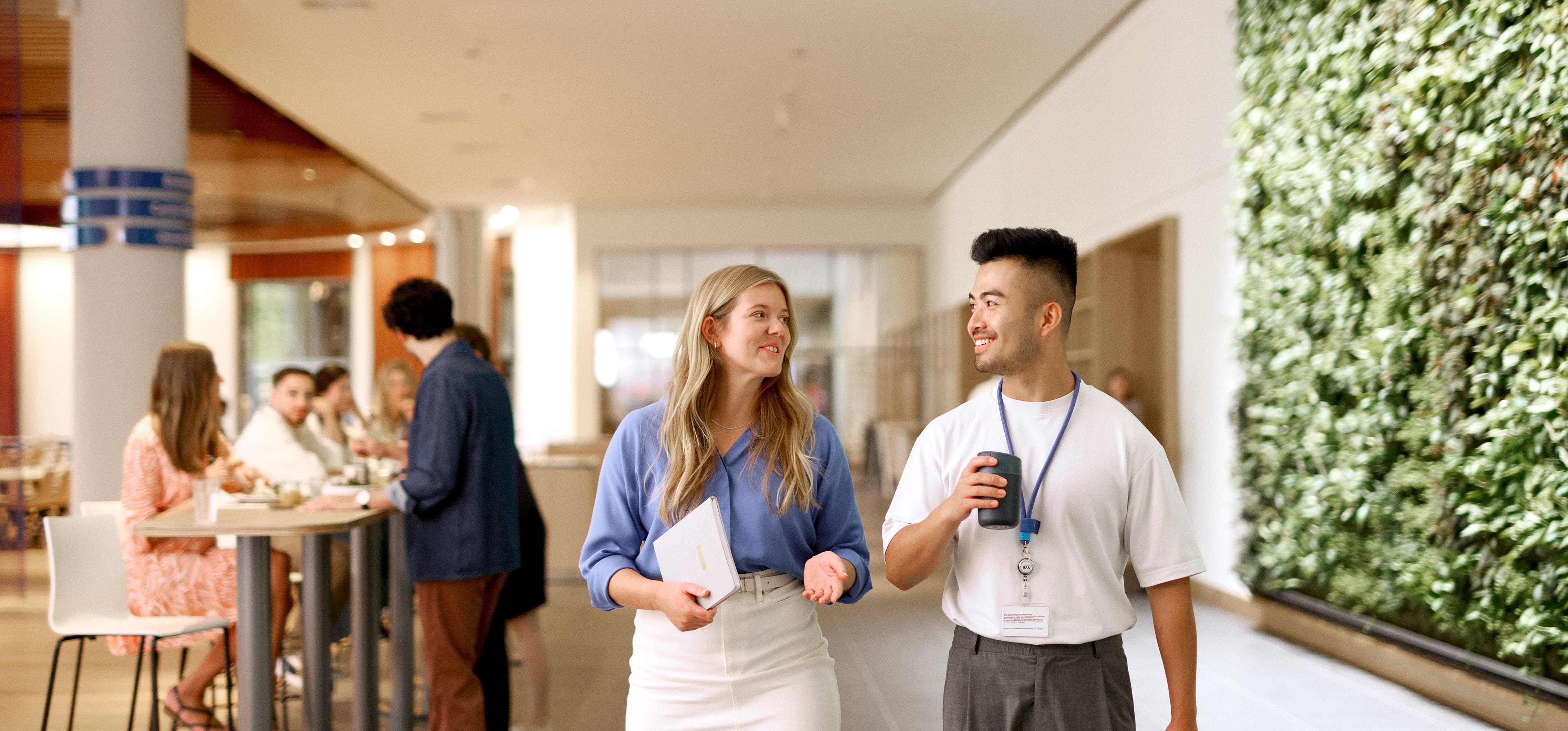 People standing and talking in a bright indoor hallway with plants on the wall, including two individuals in the foreground holding drinks and chatting while others converse in the background.