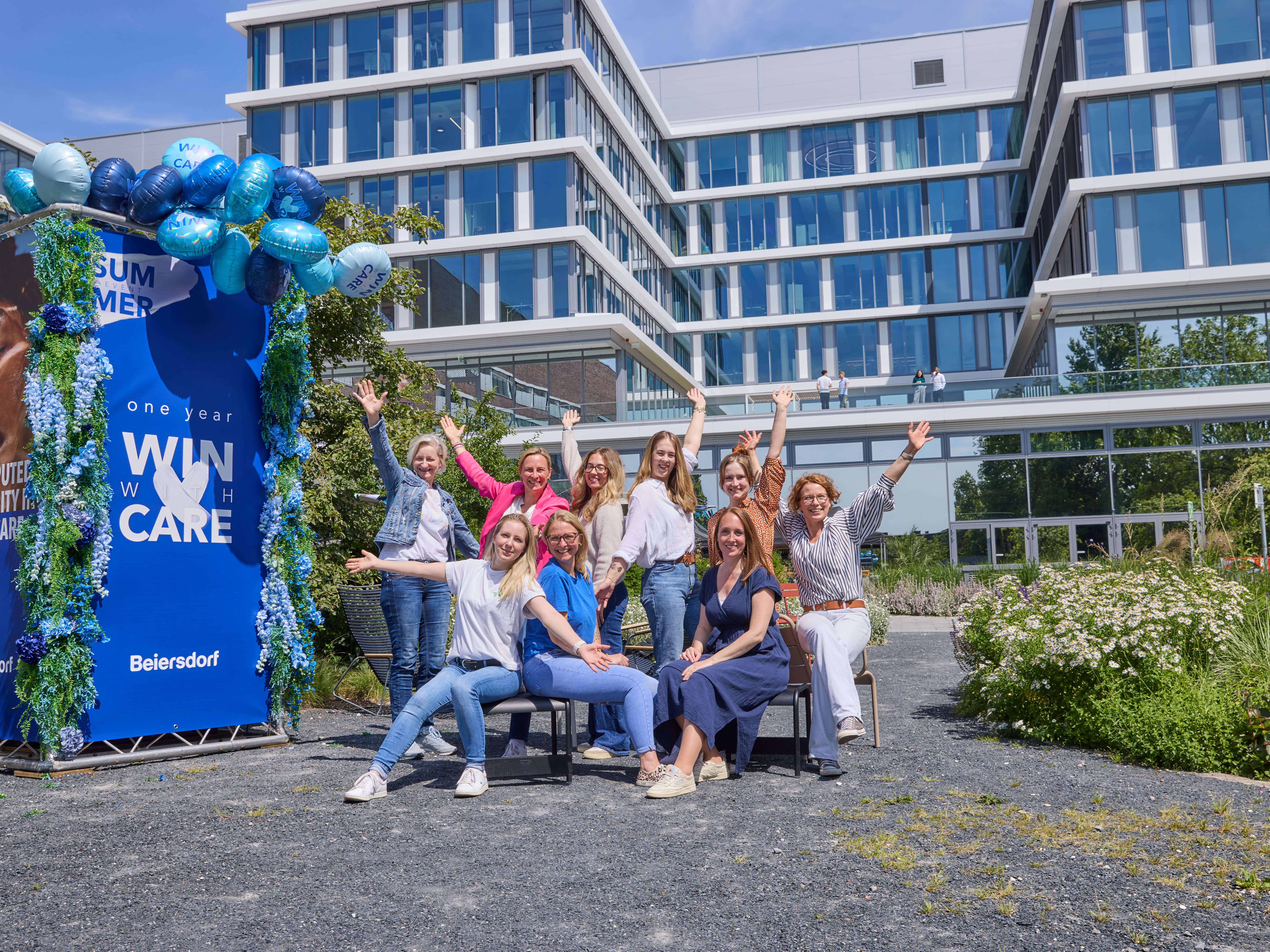 Group of people posing outdoors in front of a modern building, sitting and standing around a blue display with balloons, smiling at the camera.