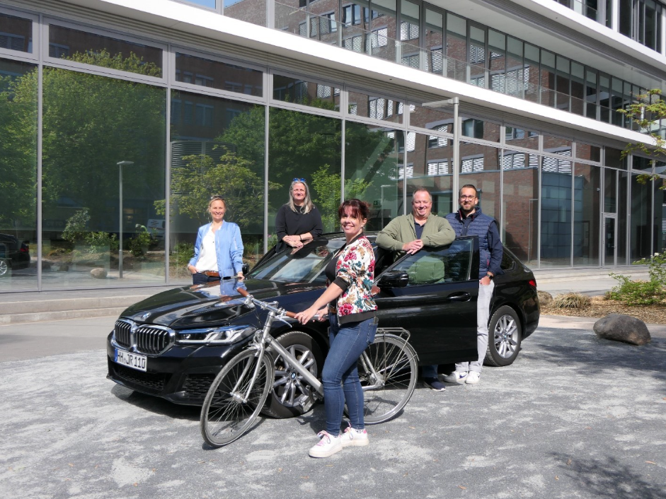 Group of people posing outdoors in front of a modern building, with one person standing beside a bicycle and others leaning against a black car.