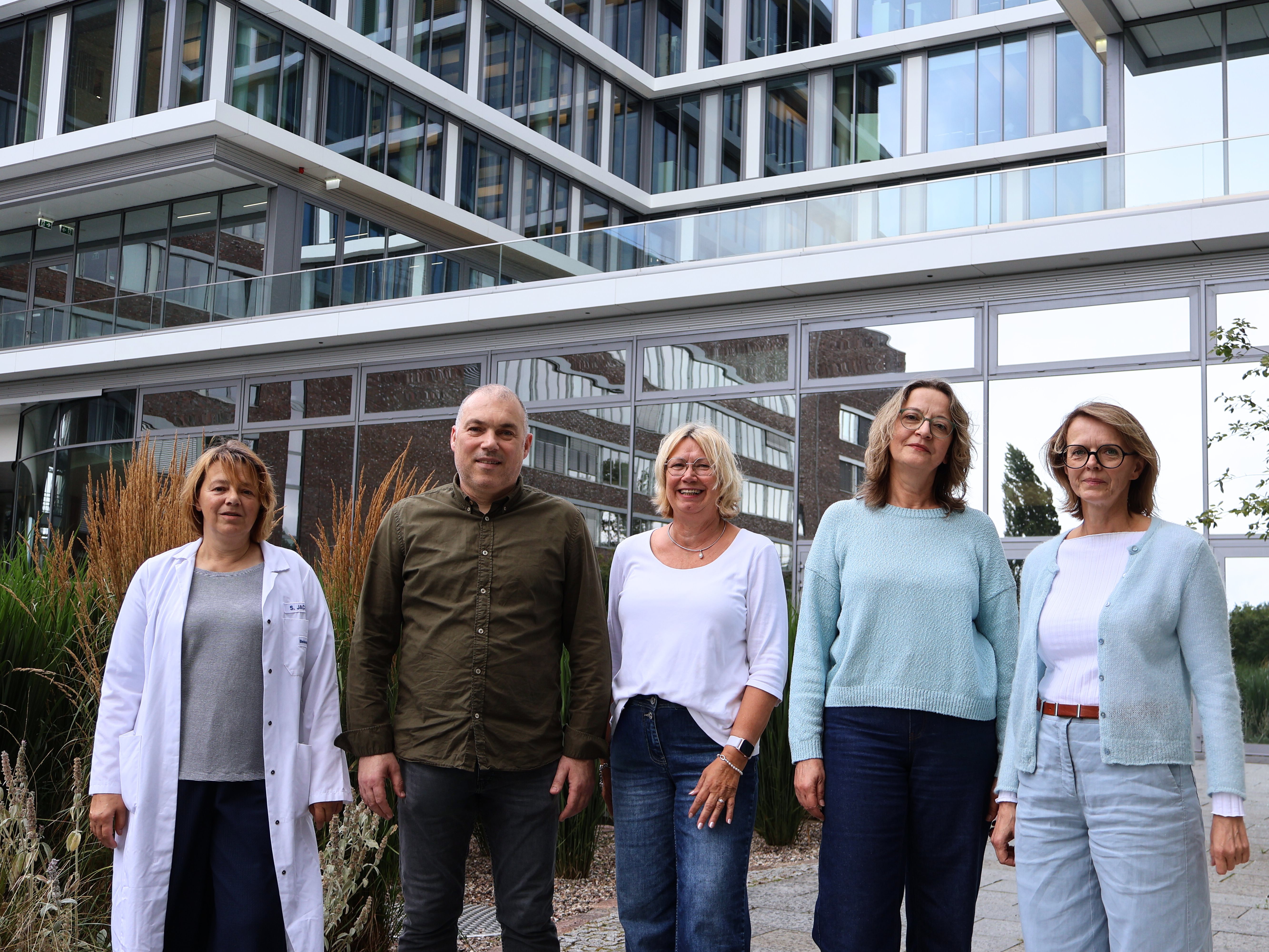 Five people standing outdoors in front of a modern building, smiling at the camera, dressed in casual and smart-casual clothing.