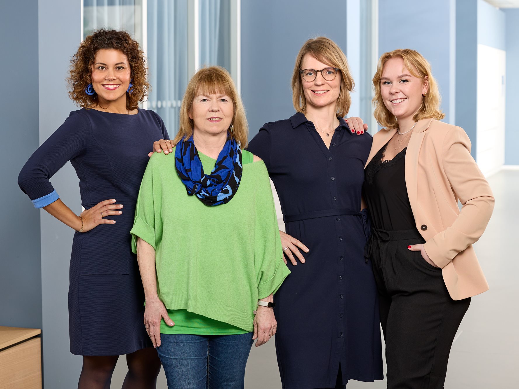 Four women standing together indoors, posing and smiling in front of tall windows, dressed in smart-casual and business attire.
