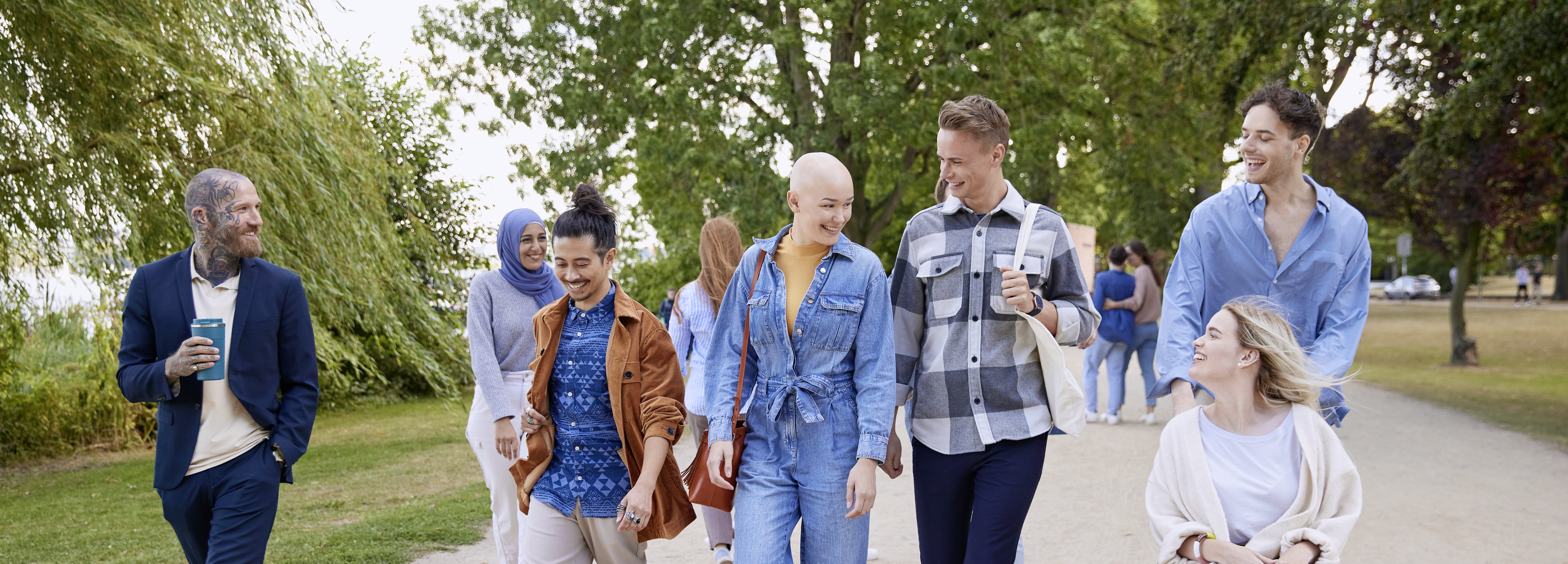 A group of people walking outdoors in a park-like setting, with a bald person in a blue denim outfit smiling while talking to others beside them.