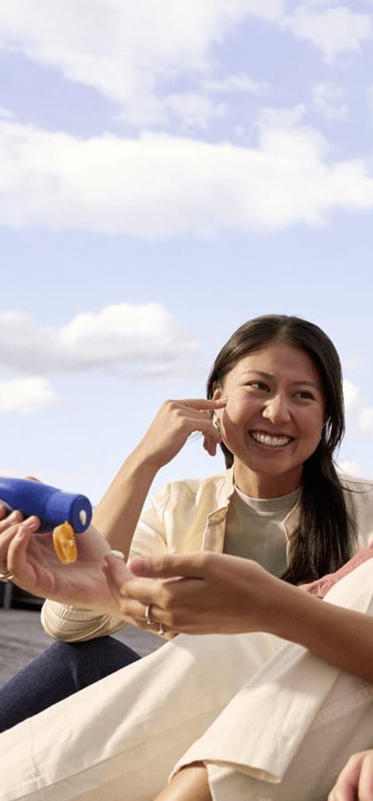 Young woman smiling while holding a sunscreen bottle outdoors, enjoying a sunny day with a relaxed expression.