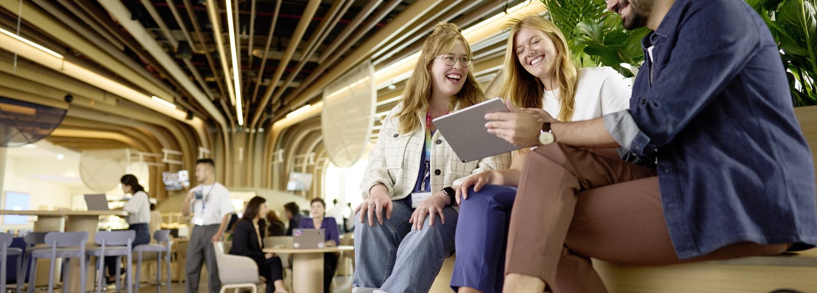 People sitting together indoors in a modern open space, smiling and looking at a laptop, with others visible in the background.