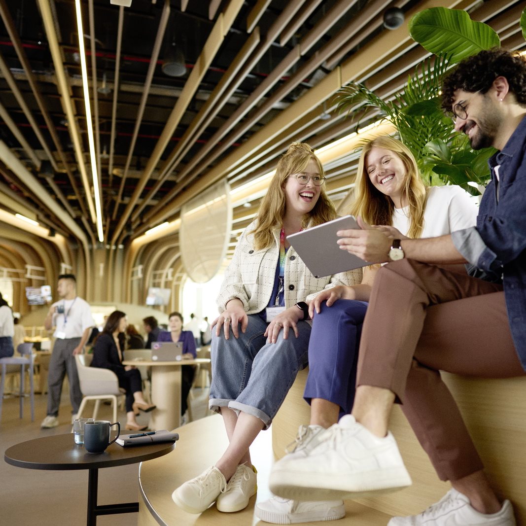 Group of young adults sitting together in a modern indoor workspace, smiling and looking at a laptop in a relaxed, collaborative setting.