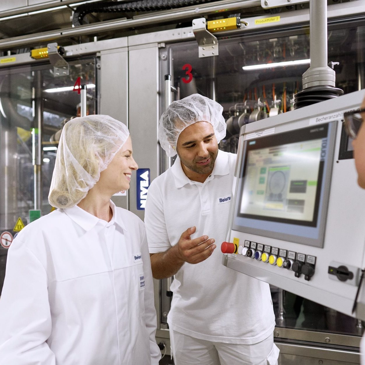Two factory workers in protective clothing and hair nets discussing information displayed on a control screen in an industrial production environment.