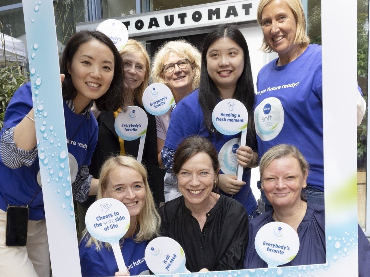 Group of smiling colleagues posing together at an outdoor event, holding branded signs and standing behind a decorative photo frame.