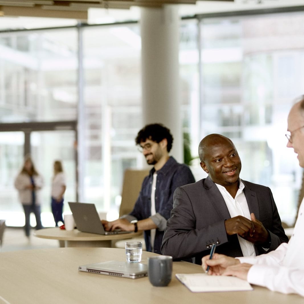 Three people sitting at a long table in a modern office or café space, talking and working, with laptops, notebooks, and drinks on the table.