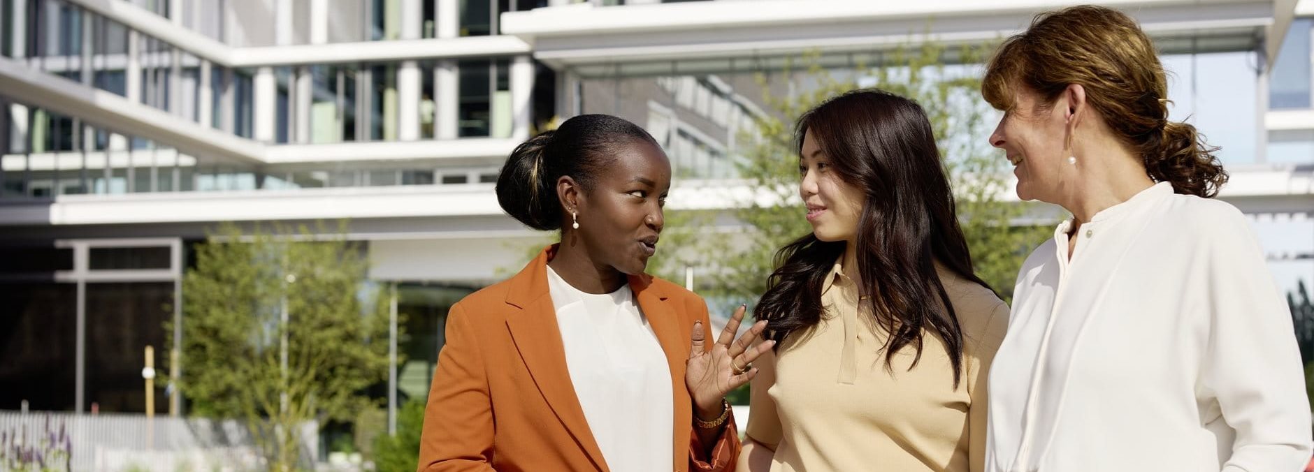 Three women standing outdoors near a modern building, talking together, with one gesturing while the others listen attentively.