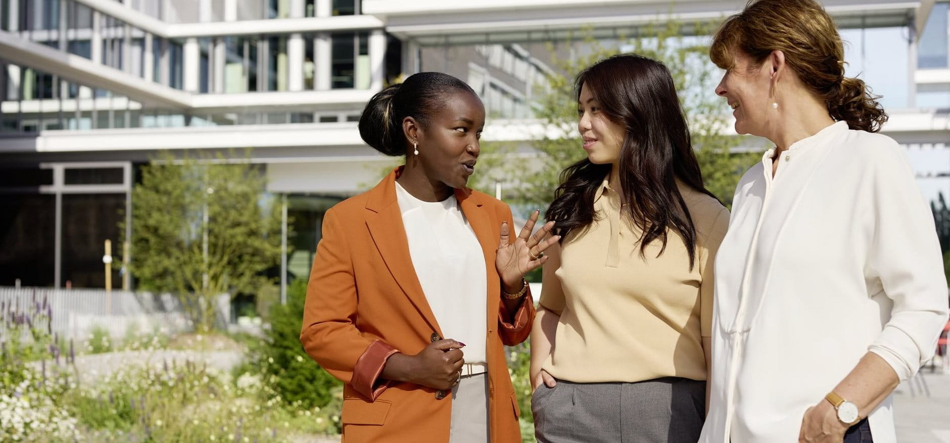 Three women standing outdoors near a modern building, talking together casually, with one holding a takeaway coffee cup.
