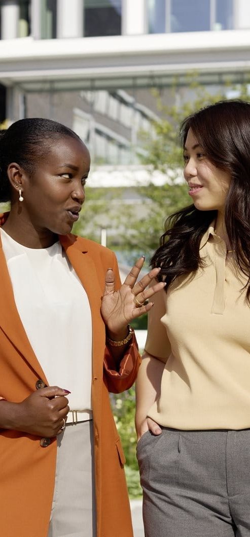 Three women standing outdoors near a modern building, engaged in conversation, with one gesturing while the others listen.