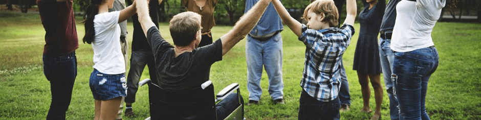 A diverse group of people standing in a circle on a grassy field, holding hands; one person using a wheelchair is included in the circle.