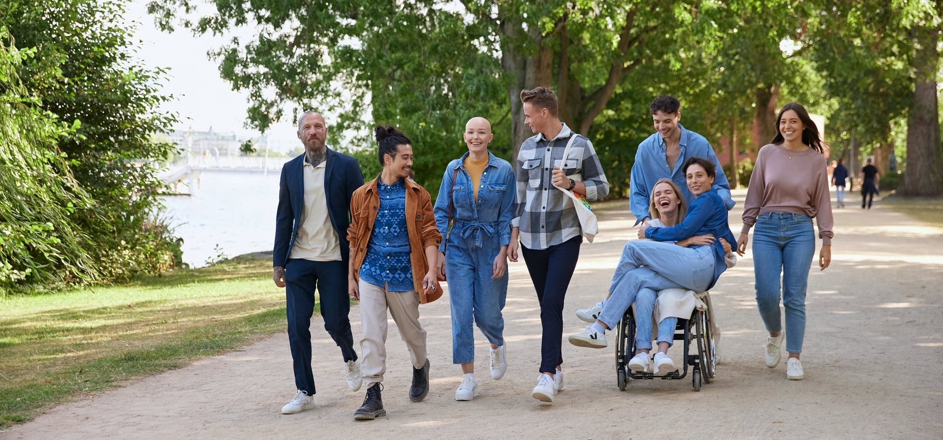 Group of people walking together outdoors along a tree-lined path, smiling and talking, with one person using a wheelchair.