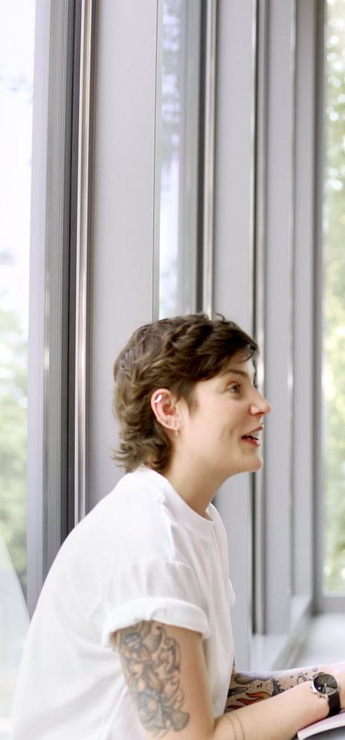 Two people sitting together in a bright office setting, smiling and talking, with plants and large windows in the background.