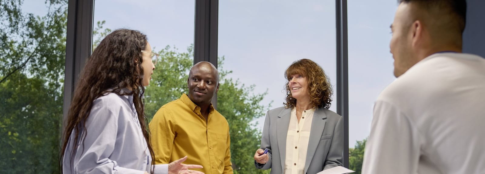 Group of people standing around a table in a bright office space, engaged in conversation during a meeting.