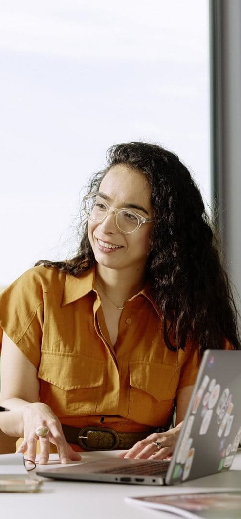 Woman smiling while working at a table with a laptop in a bright room, engaged in a relaxed professional discussion.