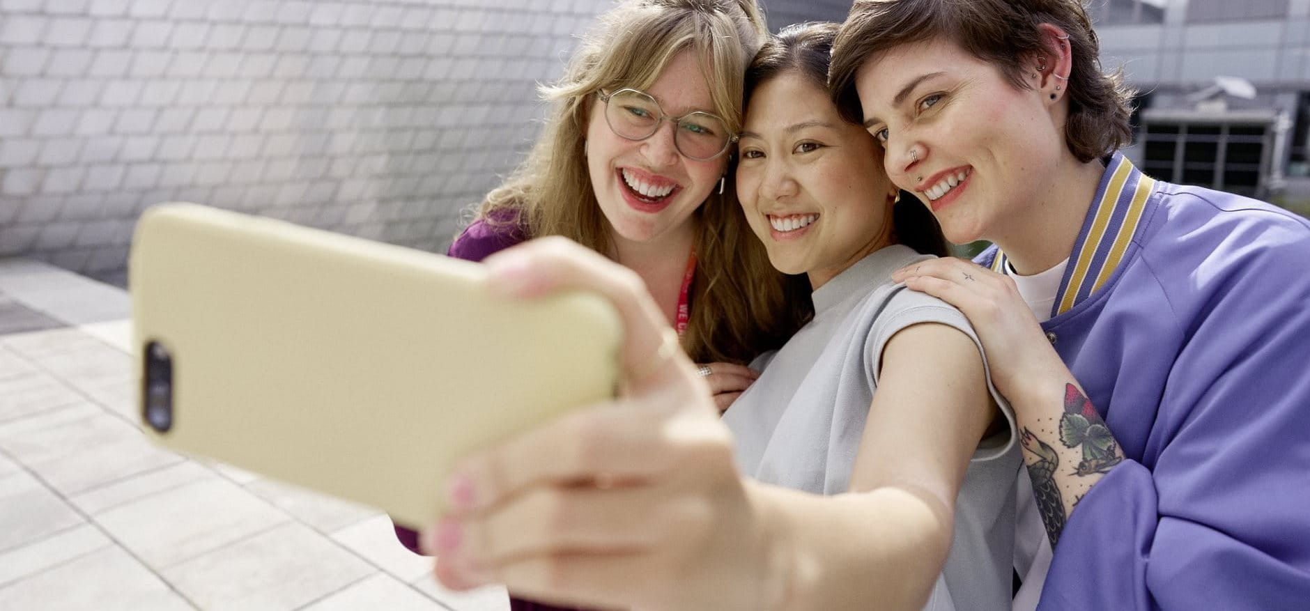 Three people smiling and taking a selfie together outdoors, holding a smartphone at arm’s length.