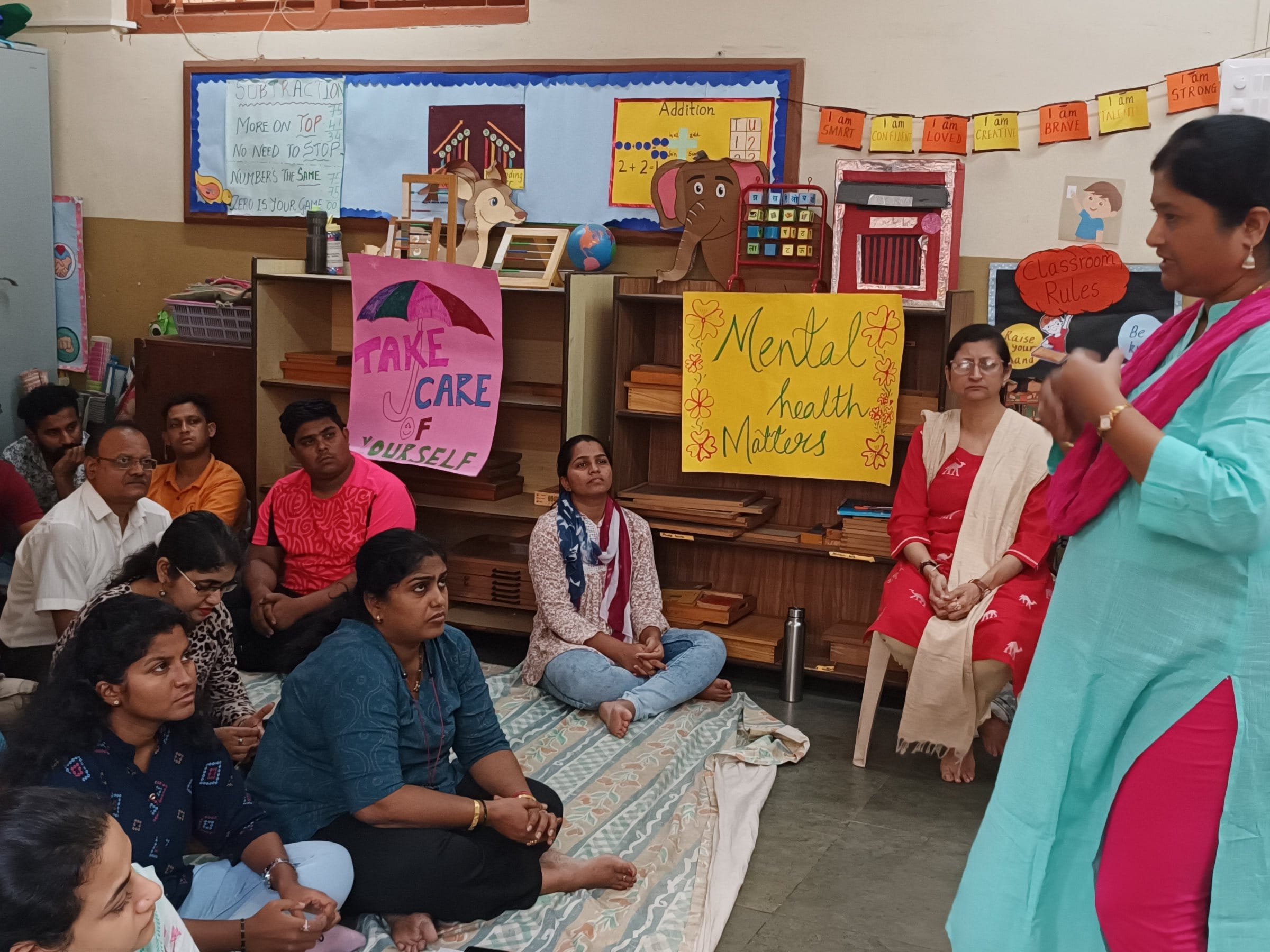 Group of girls and women sitting together in a classroom while a facilitator leads a group discussion, with posters about care, mental health, and wellbeing on the walls.