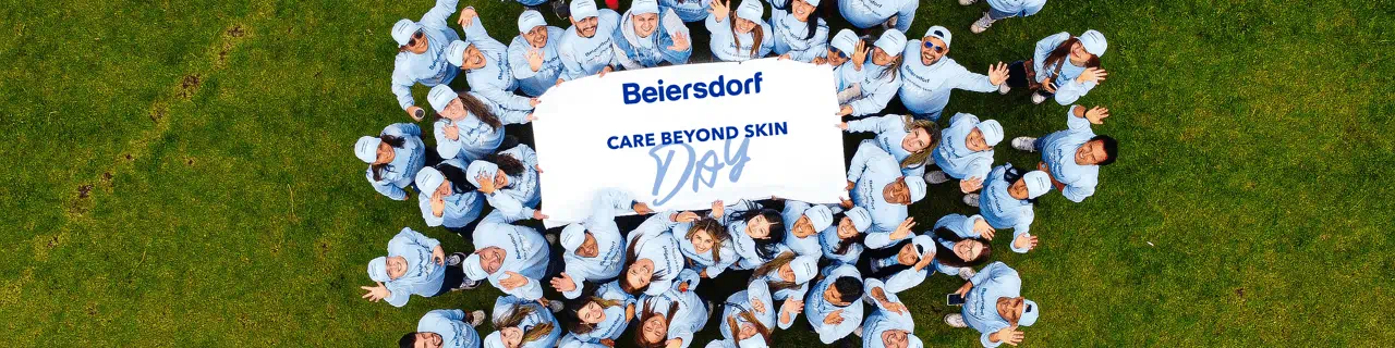 Aerial view of a large group of people in matching light-blue outfits gathered on grass, holding a banner that reads ‘Beiersdorf – Care Beyond Skin Day’ and waving up at the camera.