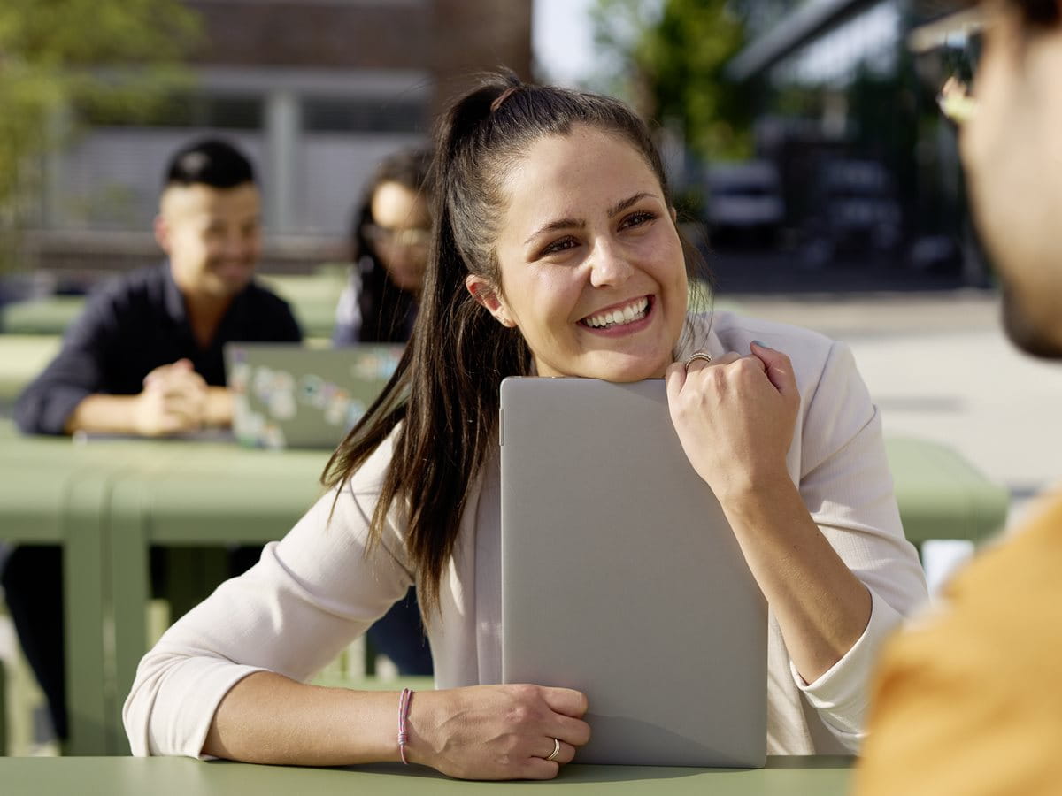 Woman smiling and talking with a colleague while holding a notebook during an outdoor discussion at a campus or courtyard table.