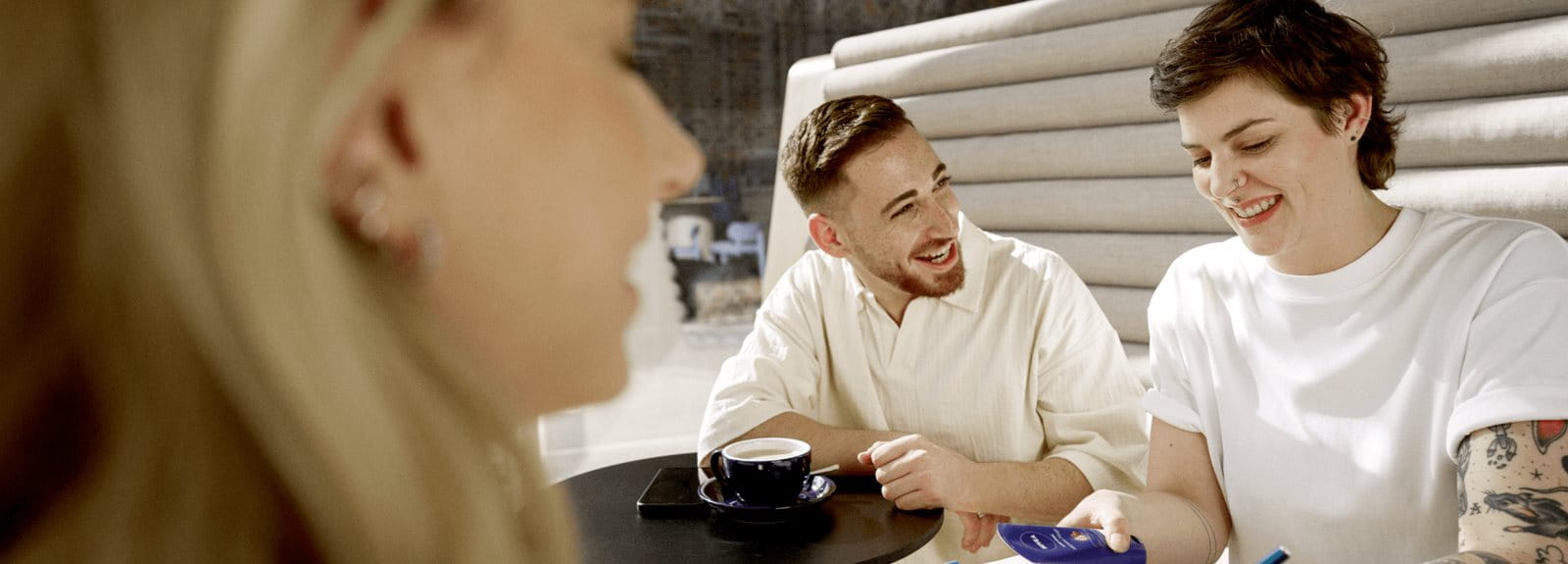 Group of young adults sitting together outdoors, smiling and chatting while sharing items at a table in a casual social setting.