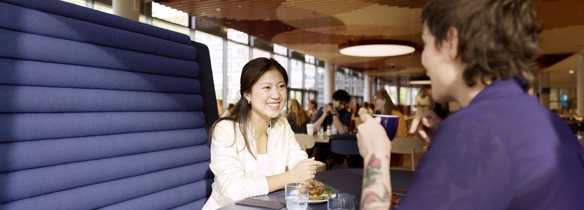 Two people sitting at a table in a busy indoor café or communal space, smiling and talking with each other.