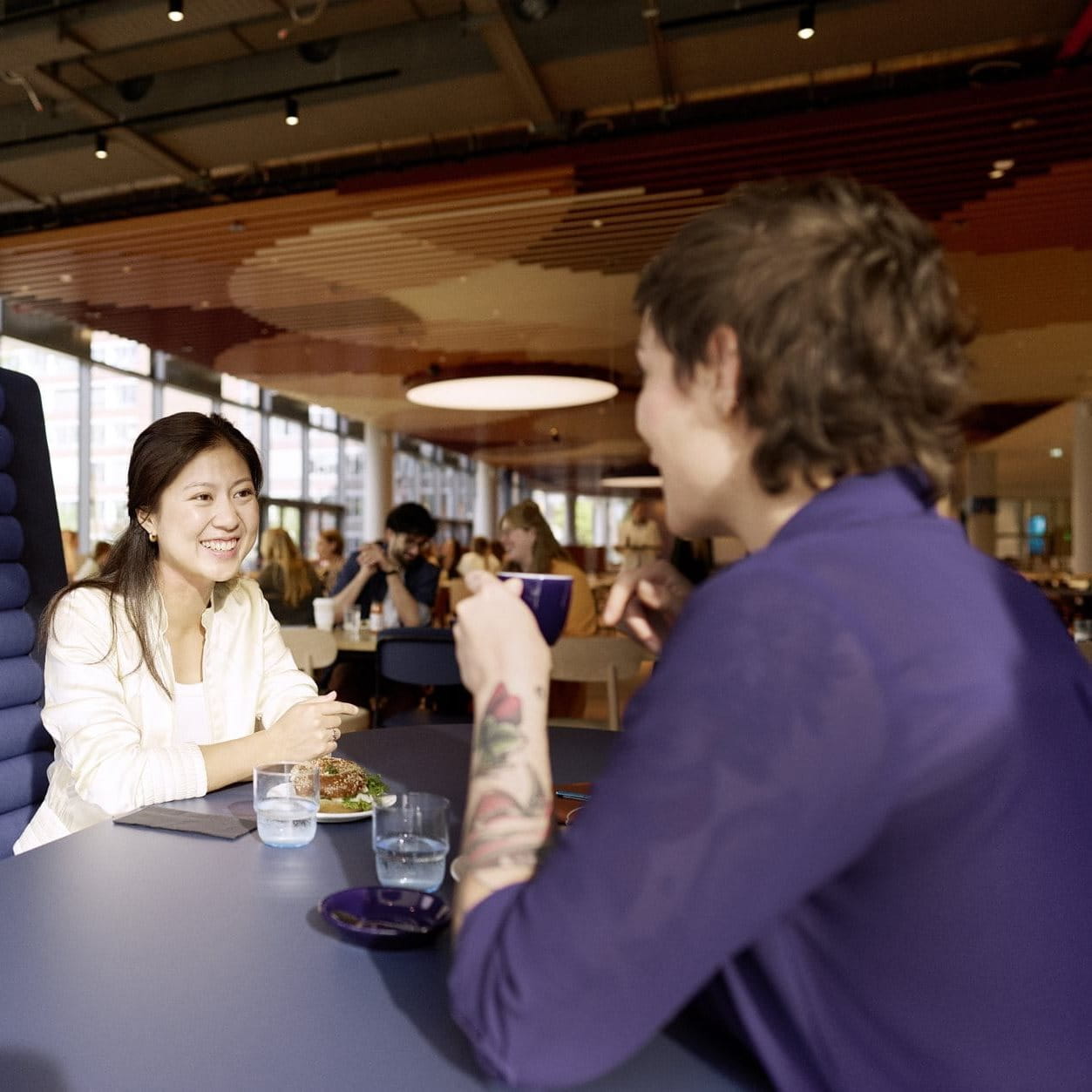 Two people seated at a table in a modern café or communal space, talking and smiling during a relaxed conversation.