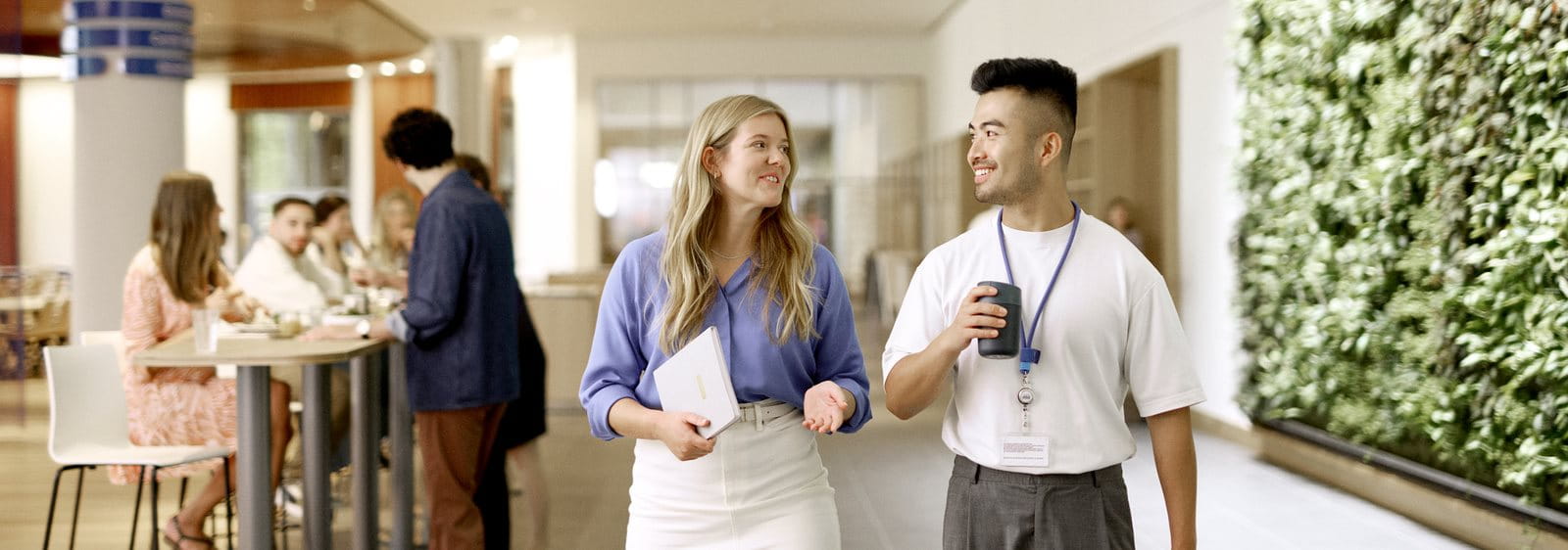 Two people walking and talking together in a bright indoor hallway, both smiling and holding drinks, with others in the background.