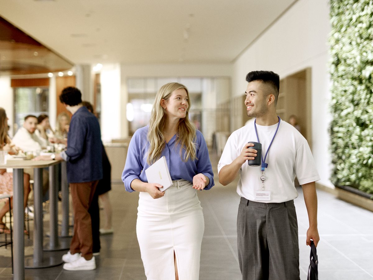 Two colleagues walking and chatting in a modern office corridor, holding a notebook and a takeaway coffee.