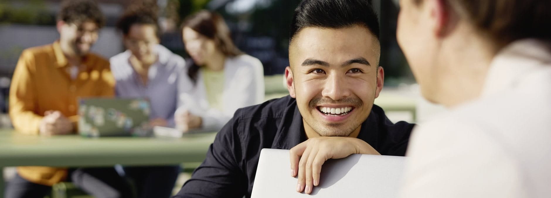 Close-up of a smiling person in a casual indoor setting, leaning forward while talking with someone off camera, with others blurred in the background.
